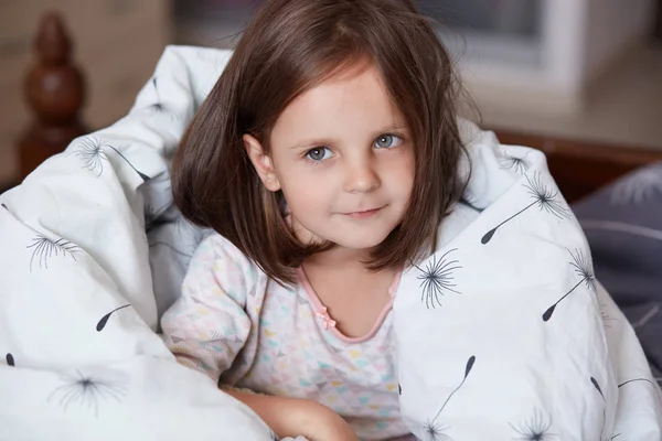 Close up portrait of sweet little dark haired girl sitting in her bed and wrapped white blanket, looking at camera and smiling while being at home in cosy room, female kid wearing white pajama. — Stock Photo, Image