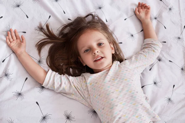 Close up portrait of cute little girl dreaming in bed, lying on white linen with dandelion and spreading arms, looking at camera, charming female kid having dark hair, resting in bed in morning. — Stock Photo, Image