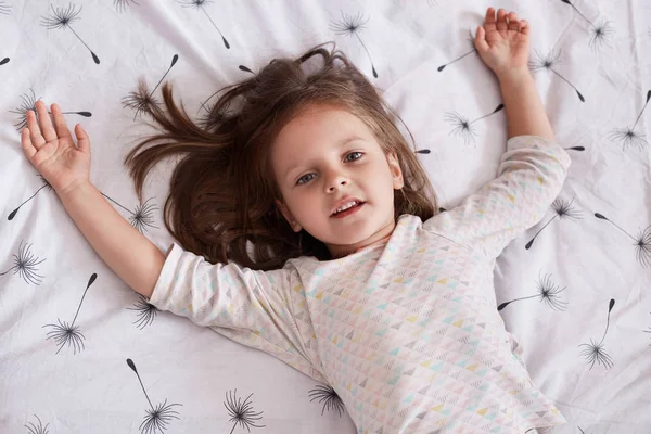 Portrait en gros plan d'une jolie petite fille rêvant au lit, allongée sur du linge blanc avec du pissenlit et des bras écartés, regardant la caméra, charmante jeune fille aux cheveux foncés, se reposant au lit le matin . — Photo