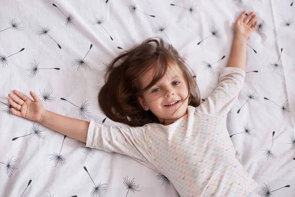 Schattig klein meisje dat thuis speelt terwijl ze op een deken ligt met paardebloem op bed met slaperige en gelukkige gezichtsuitdrukking, haar armen spreidend. Sweet elementary age kind hebben plezier in gezellige slaapkamer. — Stockfoto