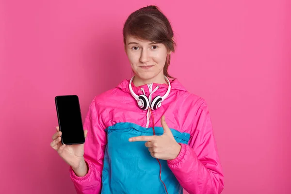 Jolie femme caucasienne souriante en chemise de sport bleue et rose montrant écran de smartphone vierge et pointant son doigt vers le téléphone, modèle posant isolé sur fond rose. Espace de copie pour la publicité . — Photo