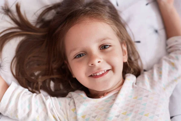 Όνειρα γλυκά. Γοητευτικό θηλυκό παιδί ξαπλωμένο στο κρεβάτι πάνω σε λευκό μαξιλάρι στο άνετο δωμάτιό της, προσπαθεί να κοιμηθεί, έχοντας χαρούμενη έκφραση προσώπου και κοιτάζει χαμογελώντας στην κάμερα, χαλαρώνοντας το πρωί. — Φωτογραφία Αρχείου
