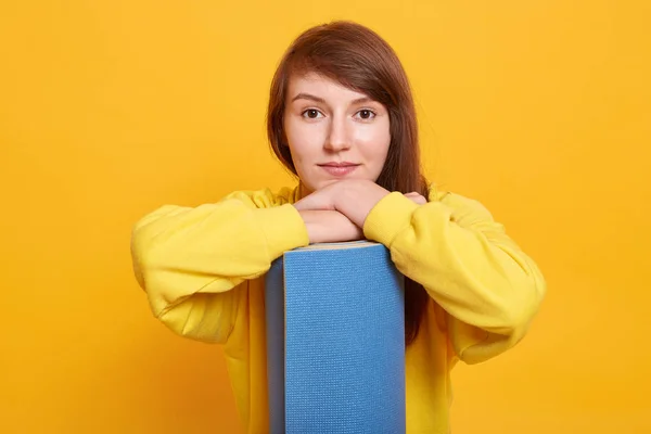 Retrato de media longitud de una joven hembra posando con una esterilla de ejercicio azul, se encuentra sonriendo aislada sobre un fondo amarillo, mirando a la cámara, lista para ejercicios deportivos, vestidos femeninos suéter con estilo . — Foto de Stock