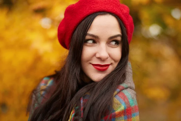 Сказочная романтичная женщина с длинными темными волосами в красном берете на размытом осеннем фоне. Девушка в лесу с оранжевыми осенними листьями, модель, завернутая в одеяло, смотрит в сторону с кокетливой улыбкой . — стоковое фото