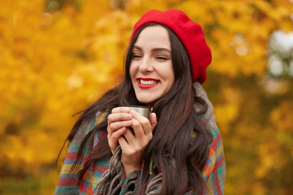 Наружный снимок красивой девушки с утренним кофе в термосе чашка, очаровательная женщина выражает счастье, женщина обернута теплым клетчатым одеялом и платья стильные красные береты, позируя в осеннем парке . — стоковое фото