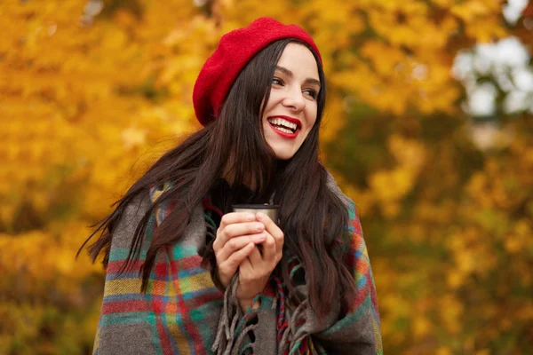 Happy woman wearing stylish red beret and wrapped warm checkered blanket, looking aside and laughing, holding thermos cup of coffee or tea, spending time in open air, enjoying autumn days in park. — Stock Photo, Image