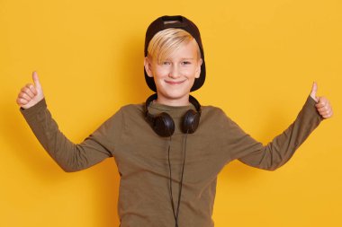 Gülümseyen ve müzik dinleyen, kulaklık takan, yeşil kapüşonlu ve siyah şapkalı küçük bir çocuğun portresi. Çocukluk konsepti..