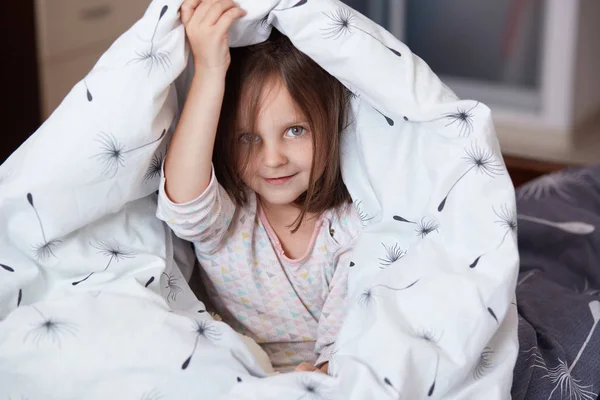 Closeup portrait of happy little girl sitting on bed under blanket and posing in room of her flat holds plaid under her head, looks directly at camera, hides fromher parents, plays at home with mother — Stock Photo, Image