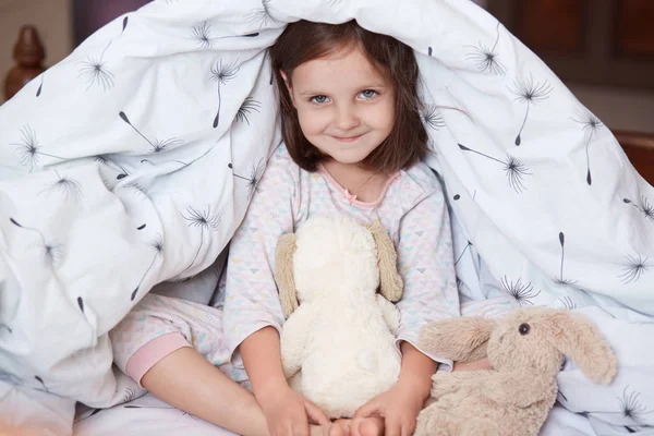 Studio shot of adorable child sitting in her bedroom with cute soft dog toy and teddy bear, relaxing in morning, playing games with her toys, looking at camera with charming smile. Childhood concept. — Stock Photo, Image