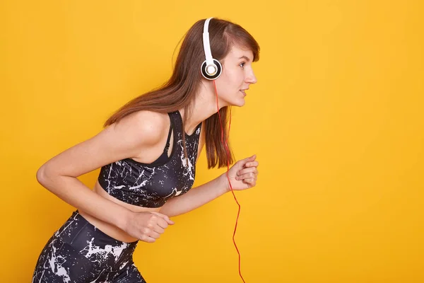 Porträt einer fitten und sportlichen jungen brünetten Frau, die sich auf das Laufen vorbereitet, Musik mit weißen Kopfhörern hört, schwarzes Top und Leggins trägt und isoliert über gelbem Studiokonzept posiert. — Stockfoto