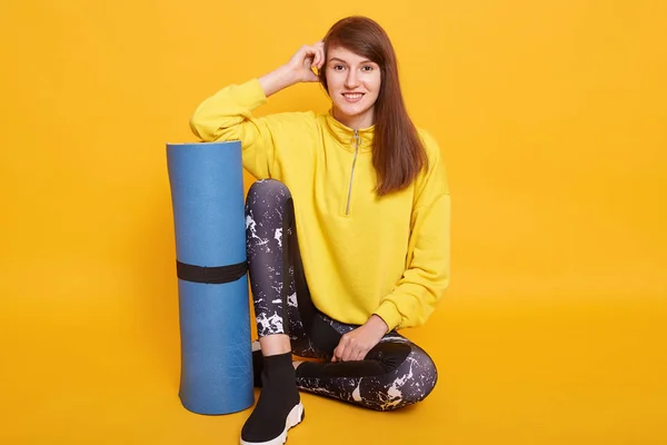 Een foto van een fitnessvrouw die met een trainingsmat zit en direct naar de camera kijkt, een schattige vrouw die traint in een fitnessclub, een stijlvolle gele capuchon en zwarte leggins draagt. Fitness- en sportconcept. — Stockfoto