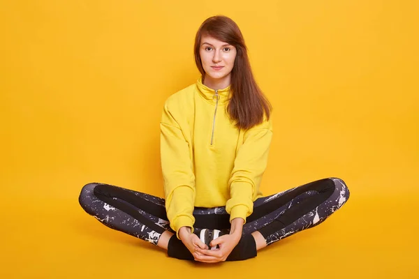 Hoprizontal tiro de mulher jovem atraente meditando enquanto sentado no estúdio floorin, posando isolado sobre fundo amarelo, feminino vestindo capuz e leggins. Esporte, fitness e estilo de vida conceito . — Fotografia de Stock