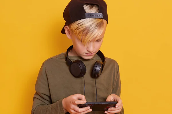 Close up of little boy using mobile phone while playing online games, cute male kid wearing gren hoody and black backwards hat keep headphones around neck. Modern technoloy concept, isolated on yellow Royalty Free Stock Images