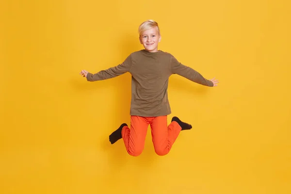 Ragazzo volante che salta con le braccia aperte alzate. Scatto completo in studio di bambino maschio con piacevole espressione facciale, posa isolata su sfondo giallo in studio. Felicità e concetto di gioia . — Foto Stock