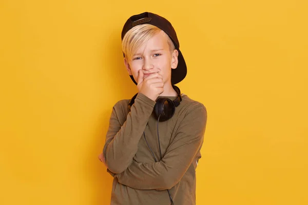 Close up portrait of little boy weraring black cap and green shirt, cute guy posing isolated over yellow background, male kid covering mouth with hands, model posing with headphones around neck.