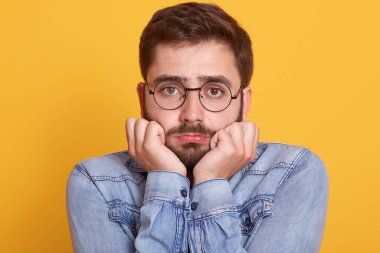 Üzgün, üzgün, sakallı, doğrudan kameraya bakan, yüzüne yakın duran, kot ceket ve gözlük takan, kötü ruh halinde olan yakışıklı bir adamın yakın portresi. İnsanlar ve duygular.