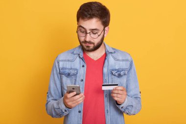 Akıllı telefonda kredi kartıyla ödeme yapan sakallı adam, konsantre görünüyor, cihazın ekranına bakıyor, cep telefonu ve kardin eli, kot ceket ve kırmızı gömlek giyiyor, sarı duvara karşı poz veriyor.