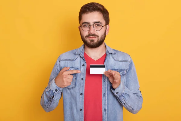 有自信的大胡子男人用食指指着信用卡 男人看相机 用信用卡购物 穿牛仔夹克和红色休闲衫的画像 — 图库照片