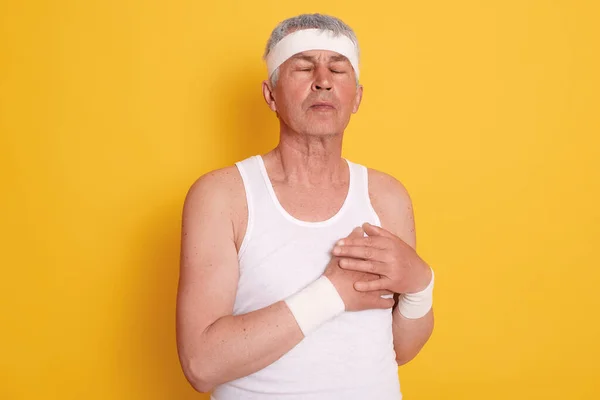 Mature man posing against yellow wall with closed eyes and touching his chest, feels pain in heart, needs treatment, has heart attack, needs first aid.