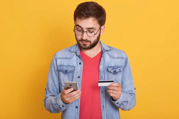 留着胡子的男人拿着信用卡在智能手机上付着钱 看上去很专注 看着手机的屏幕 手牵着手机和卡丁鱼 身穿斜纹棉布夹克和红色衬衫 在黄色的墙上摆出一副姿势 — 图库照片