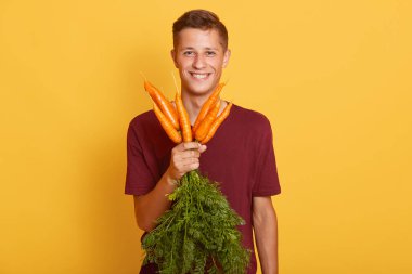 Normal giyinmiş gülümseyen genç bir adam, bir demet çiğ havuç tutan, sarı arka planda izole edilmiş poz veren, kameraya gülümseyen, çiftlikten hasat yapan bir erkek..