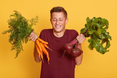 Elinde taze havuç ve pancar tutan mutlu bir erkek, tatmin olmuş görünüyor, gözlerini kapalı tutuyor, sarı duvara yaslanmış, bordo tişört giyen bir erkek, vejetaryen hasat yapmaktan memnun..
