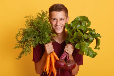 Çiftlikte çalışan mutlu genç çiftçinin portresi, hasatını gösteriyor, havuç ve pancar tutuyor, sarı arka planda soyutlanmış, gelişigüzel giyinen erkek.