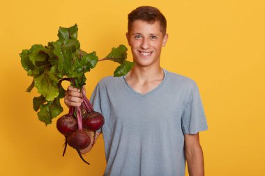 Gülümseyen genç çiftçi, elinde pancar kökleri, doğrudan kameraya bakıyor, gündelik giysiler giyiyor, sarı duvara karşı duruyor..