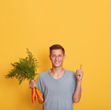 Mutlu çekici erkek işaret parmağı yukarı, reklam ya da promosyon için fotokopi alanı gösteriyor, havuçları elinde tutuyor, erkek hasat topluyor, sarı arka planda izole edilmiş poz veriyor.