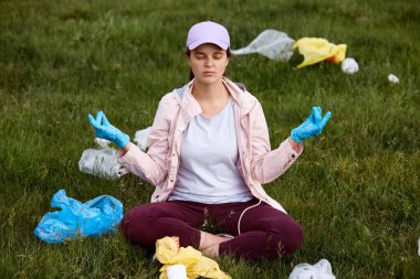 Genç kız sahada çamura saplanmış, sakinleşmeye çalışıyor, meditasyon yapıyor, çok üzgün, çöplerle çevrili, gözlerini kapalı tutuyor, ekolojik sorunlardan dolayı üzgün..