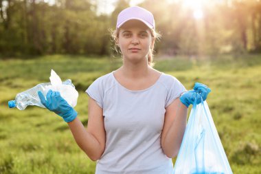 Beyzbol şapkalı, beyaz tişörtlü, mavi lateks eldivenli, çöp torbası dolu bir kadın yorgun görünüyor, kirli çayırları temizliyor, çöp topluyor. Ekolojik sorunlar.