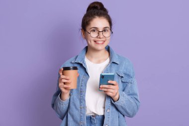 Kot ceket, beyaz gömlek ve yuvarlak gözlük giyen gülümseyen kadın elinde kahve ve cep telefonu tutan genç kız leylak arka planda yalnız poz veren kadın..