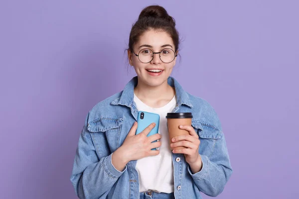 一个面带微笑的高加索女人的画像 她靠着紫丁香墙站着 端着咖啡 直接看着相机 女人们喝咖啡休息 手里拿着智能手机 检查社交网络 — 图库照片