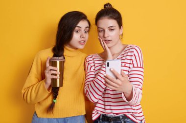 Ellerinde akıllı telefon olan ve kahveyi götüren kadınlar şok olmuş yüz ifadesiyle cihaz ekranına bakan kadınlar, şok edici haberler okuyan, sarı duvarın karşısında duran kadınlar..