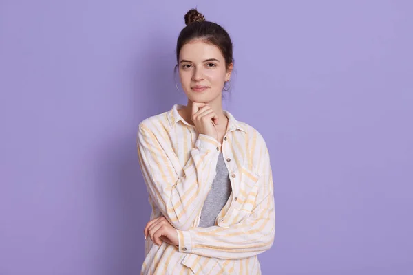 Attractive female looking at camera with hand under chin. Adorable model posing against lilac background, wearing casual attire, stands with hair bun.