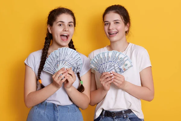 Excited females wearing white t shirts and jeans, ladies holding fans of money, keeping mouths opened, girls win lottery, look happy, celebrating winning.