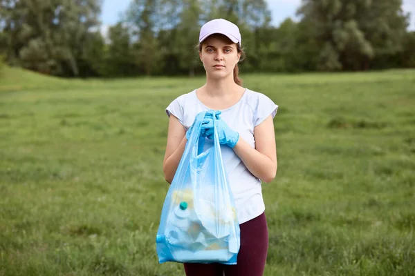 Young woman in white t shirt and baseball cap holding blue garbage bag in hands, female collecting litter in meadow, looks at camera, stands against trees on background.