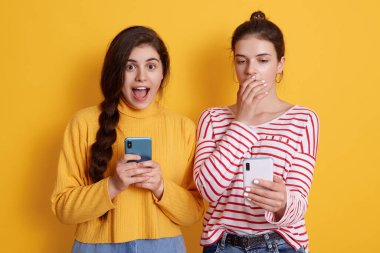 Telefonları olan iki arkadaş sosyal ağda şok edici haberler okuyorlar. At kuyruklu kadın kameraya büyük gözlerle bakıyor ve bağırıyor. Çizgili tişörtlü kadın ağzını avuç içiyle kapatıyor..