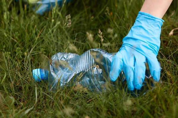 Hand of unrecognizable person in blue latex glove picks up plastic bottle from the ground, unknown person cleans up in park, meadow or field, solves ecological problems.