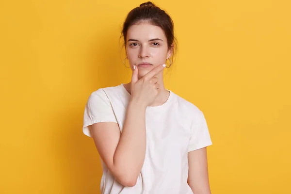 自信のある表情でカメラを見て 白いTシャツを着て深刻な若い女性は 思慮深く見える 問題を解決するか 答えの質問を見つけると思う 黄色の壁に対して立つ — ストック写真