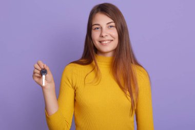 Leylak rengi duvarın karşısında duran büyüleyici gülümsemeli kadın, araba anahtarı, uzun güzel saçlı, sarı günlük gömlekli kadın, anlamlı mutluluk, yeni bir otomobil kazanıyor..