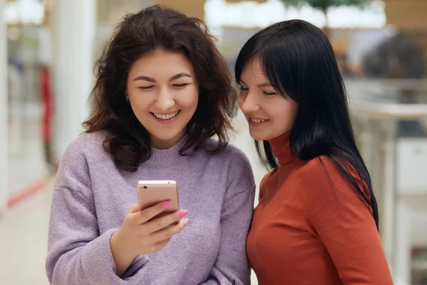 Friends having fun with smart phone, looking at mobile phones screen with happy expression and laughing, females wearing sweaters, posing together while reading social network.