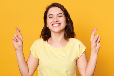 Hayallerin gerçek olduğuna inanan çekici bayan, umut dolu koyu saçlı kadın kendini şanslı hissediyor, parmaklarını çapraz yapıyor, dilek tutuyor ve büyük umutlar kuruyor, kameraya gülümsüyor, kapalı alanda poz veriyor..