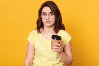 Sarı arka planda izole edilmiş kahve içen genç, güzel bir kadın korkmuş ya da şok olmuş görünüyor, koyu renk saçlı, gözlüklü ve günlük tişörtlü bir kız..