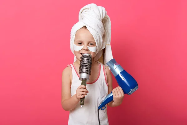 Cute female child imagines she super star and singing with comb in hands ,charming little girl with patches under eyes and towel against rosy wall, holding hairdryer in other hand.
