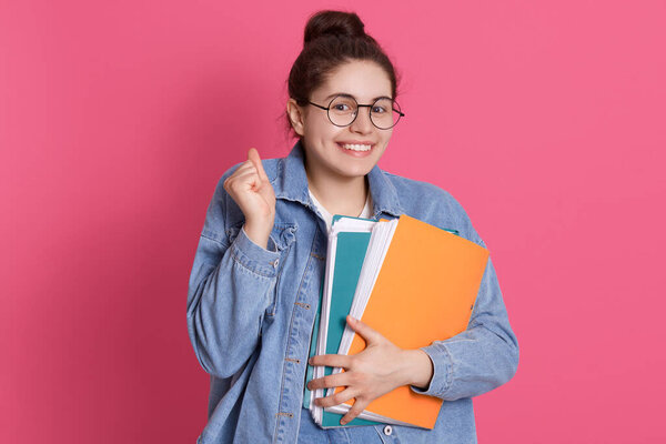 Young smiling student in eyeglasses and denim jacket standing with paper folders isolated over rosy background, clenching fist, looking smiling at camera.