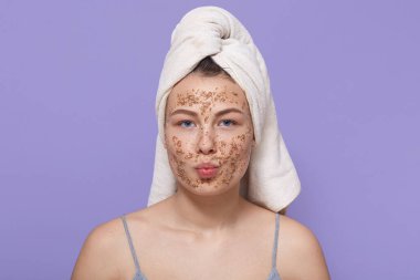 Yüzünde maske olan güzel bir kadın, dudaklarını yuvarlak tutuyor, kafasında beyaz havluyla poz veriyor, spa salonunda dinleniyor, leylak duvarda izole edilmiş, spa ameliyatlarından hoşlanan bir kadın..