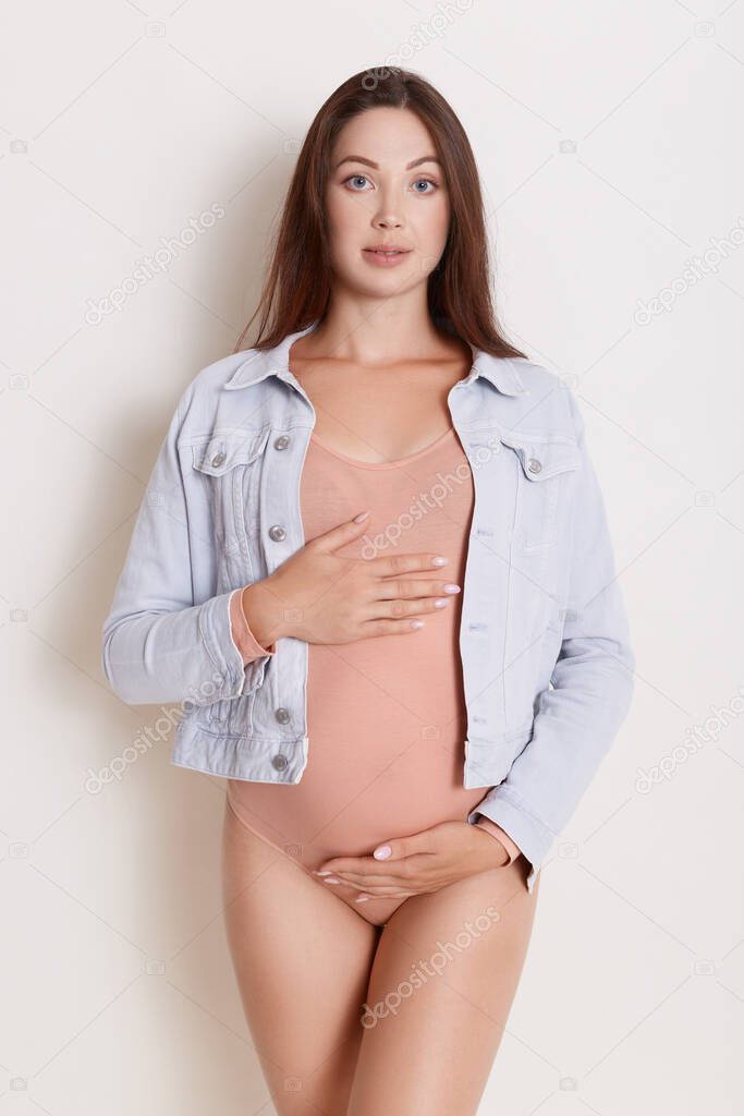 Pregnant Caucasian woman isolated over white background keeping her hands on belly, wearing pale pink bodysuit and denim jacket, looking at camera, looks calm.