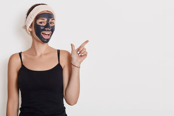 긍정적 여성은 얼굴에 영양이 마스크를 쓰는데 공간에 손가락을 옆으로 향하게 — 스톡 사진