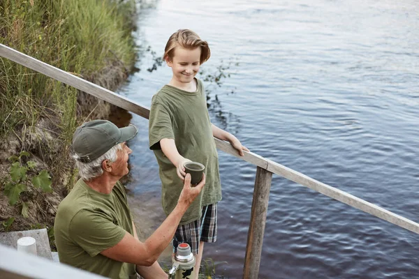 快乐的家庭一边喝热水瓶里的茶 一边欣赏钓鱼 祖父和孙子躺在通向水的木楼梯上 父亲和儿子穿着绿色的T恤衫 — 图库照片